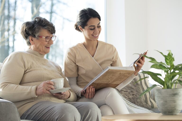 Caregiver in a beige top sharing a photo album with a smiling older woman holding a cup of tea on a sofa