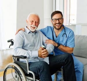 Older man in a wheelchair smiling with a friendly doctor in blue scrubs beside him.